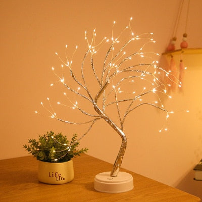 A decorative LED mini tree lamp with silver and gold colors, designed to resemble a Christmas tree, placed on a white base, with a blurred background suggesting indoor setting.
